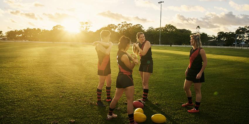 This image depicts four women on a grassy sports field during sunset, dressed in athletic uniforms. They are wearing sleeveless black tops with red diagonal stripes, black shorts, and matching socks, suggesting they may be part of a sports team, possibly for Australian rules football. The women appear relaxed and engaged in conversation, with a few footballs lying on the ground nearby.