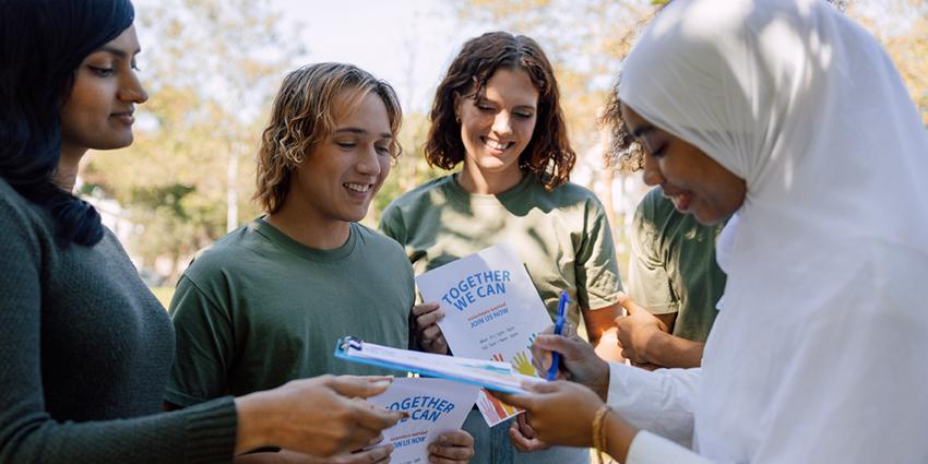 This image features a group of young people gathered outdoors, engaging in a charitable or community event. One person, dressed in a white headscarf and attire, is signing a document on a clipboard held by someone else. The others are smiling and holding flyers that read "TOGETHER WE CAN" with the subtitle "Join Us Now!"