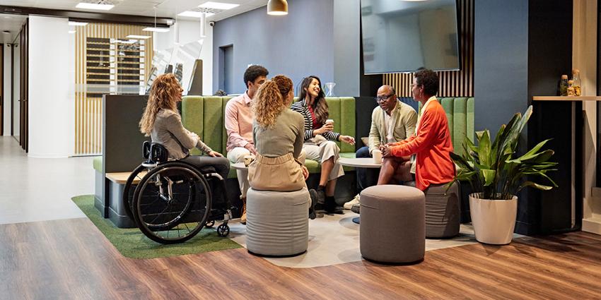 This image shows a diverse group of people engaged in a discussion in a modern, open office setting. They are seated in a semi-circle on various seats, including a wheelchair, stools, and a built-in bench with green upholstery. The group appears to be having a collaborative conversation, with everyone engaged and facing each other, creating an inclusive and supportive atmosphere.