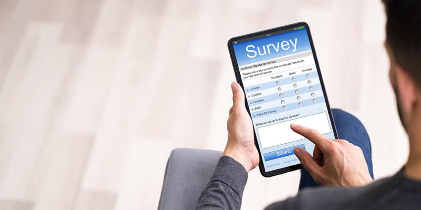 This image depicts a person holding a tablet displaying an online survey form. The survey appears to be about customer satisfaction, with various questions on aspects like location, comfort, fitting, and staff, each with a rating scale from "Excellent" to "Average." There’s also a text box at the bottom labeled "How can we improve our service?" where respondents can add additional feedback.