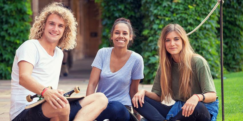 Three young adults are seated outdoors on a grassy area, smiling at the camera. They are dressed casually, and one person holds a skateboard, suggesting a relaxed, youthful atmosphere. 